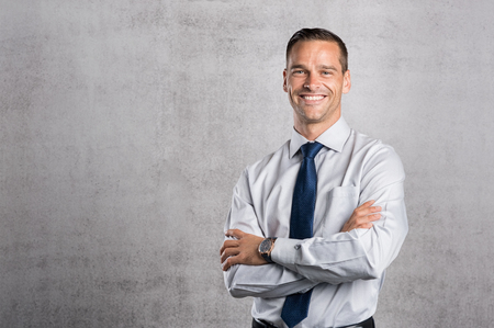 Happy businessman looking at camera on grey background with copy space. Handsome young business man standing against a grey wall with crossed arms. Successful formal man smiling with copy space.の写真素材