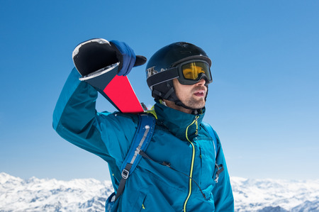 Pensive skier carrying ski on shoulders on the peak of the snowy mountain. Portrait of young thoughtful man looking snowy panorama before ski. Holiday and winter sport concept.の写真素材