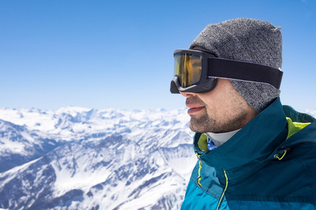 Portrait of man wearing ski glasses and cap while looking the snowy mountains. Closeup face of sporty guy contemplate the snowy. Satisfied skier smiling and wearing ski mask in mountains.の写真素材