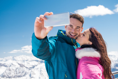 Smiling couple taking selfie with smartphone with snowy mountains in background. Young couple enjoying vacation during winter season outdoor. Girlfiend kissing boyfriend on cheek while man taking selfie.の写真素材