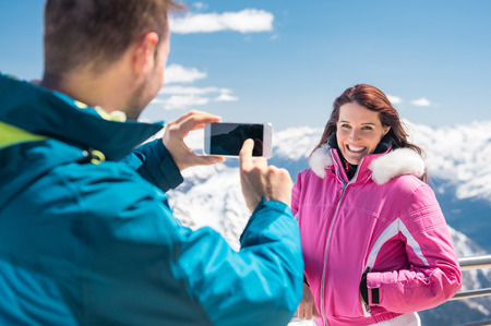 Boyfriend clicking a picture with smartphone of a girlfriend with snowy mountains in background.の写真素材