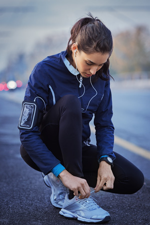Young woman athlete with earphones listening to music and tying laces on street.の写真素材