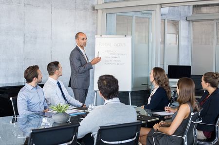 Confident mature businessman giving a presentation to his team in modern office. Business brief with annual goals with employees. Leadership man training businessmen and businesswomen in conference room.の写真素材