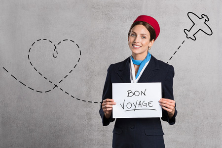 Young air hostess holding Bon Voyage sign isolated on grey background. Flight assistant holding sign with airplane route in the background. Woman stewardess with board and copy space.の写真素材