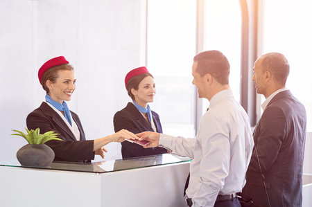 Airline passengers checking in at airline counter. Young woman giving passport and ticket back to businessman at airport check in. Two businessmen at airport checkin desk leaving on business trip.の写真素材