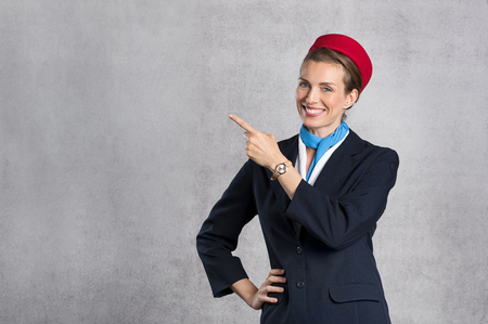 Portrait of air hostess pointing against grey background with copy space. Happy flight assistant indicating. Young flight attendant on gray background showing something while looking at camera.の写真素材