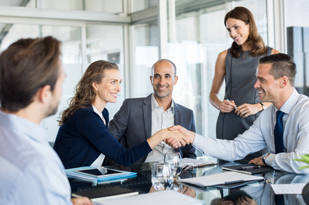 Handshake to seal a deal after a meeting. Two successful business people shaking hands in front of their colleagues. Mature businesswoman shaking hands to seal a deal with smiling businessman.の写真素材