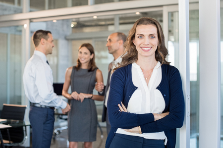 Portrait of executive businesswoman standing in office while team working in background. Smiling business woman leaning against glass wall in meeting and looking at camera. Successful mature woman at work.の写真素材