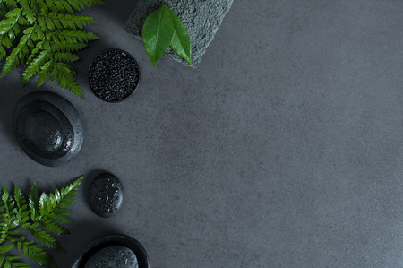 Top view of spa setting with wet hot stones and green ferns on gray background with copy sapce. High angle view of stacked black stones for massage with drops of water and grey rolled towel on blackboard. Luxury and elegant wellness concept.の写真素材