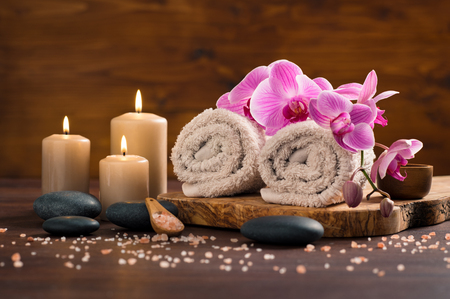 Spa setting with brown rolled towel, orchids and candles on wood. Relaxing spa concept with candles, towels and hot stones massage with himalayan pink salt. Beautiful composition for beauty treatment in a spa.の写真素材
