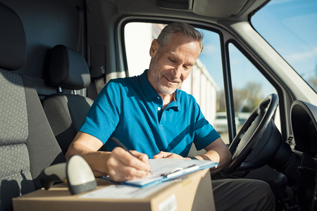 Happy delivery man with cardboard box checking list in van. Smiling courier checking list on clipboard. Deliveryman sitting in van and checking the delivery list.の写真素材