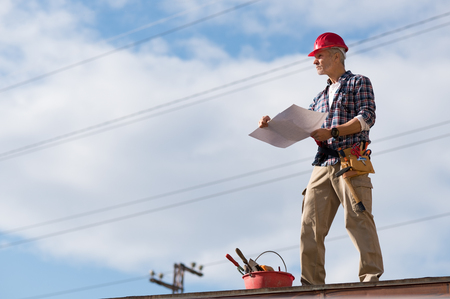 Engineer standing on rooftop looking away and holding blueprint project. Mature construction worker kit standing on rooftop with copy space. Workman inspecting construction site with sheet of paper in hand.の写真素材