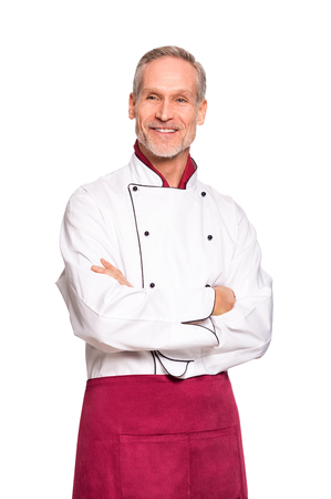 Confident professional chef with crossed hands isolated on white background. Happy mature cook with red apron and white uniform. Portrait of satisfied chef man with crossed arm looking away.の写真素材