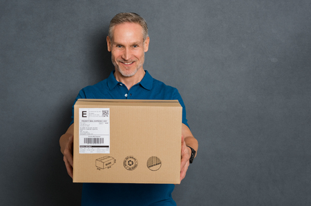 Happy delivery man stretching out his arms to give the package. Smiling courier giving card box on grey background. Portrait of deliveryman handing parcel to customer isolated with copy space.の写真素材