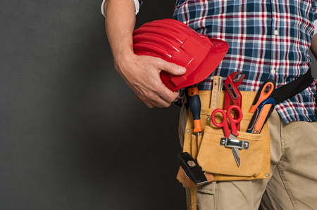 Closeup of hardhat held by construction worker on grey background. Detail of bricklayer holding red helmet and kit tool. Closeup of craftsman hand holding tool belt with equipment on grey wall with copy space.の写真素材