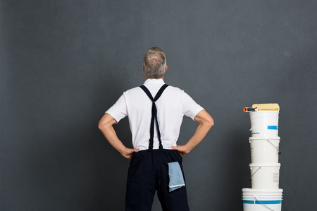 Rear view of painter looking at grey wall to paint. Mature workman with paint bucket and roller looking up isolated on gray background. Craftsman looking at blank wall with copy space.の写真素材