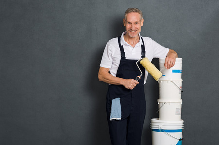 Smiling painter holding a paint roller and leaning on stack of paint bucket. Portrait of happy mature painter looking at camera isolated over grey wall. Satisfied handyman with roller and paint bucket with copy space.の写真素材