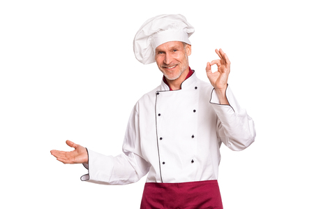 Smiling senior cook showing something isolated on white background. Portrait of happy mature chef presenting over white background. Cheerful chef man looking at camera isolated on white with hand sign ok.の写真素材