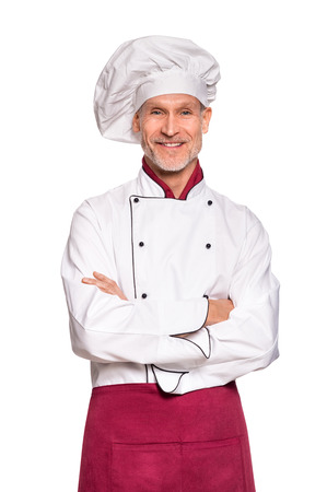 Restaurant experienced chef standing with folded arms isolated on white background. Professional cook standing in white uniform and red apron. Portrait of happy and successful chef with cap looking at camera.の写真素材