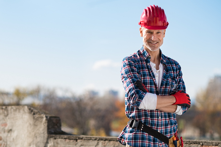 Smiling craftsman standing with arms folded and looking at camera. Happy construction worker with tool kit on waist and work gloves standing on rooftop. Portrait of satisfied bricklayer with copy space.の写真素材