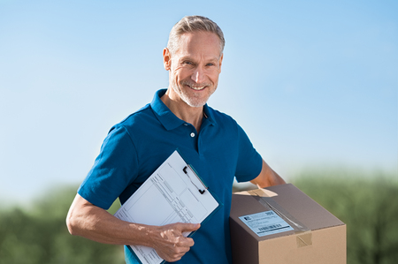 Smiling delivery man holding package and bill. Postman in blue uniform holding parcel and looking at camera. Happy courier man holding cardboard box and clipboard with delivery receipt.の写真素材
