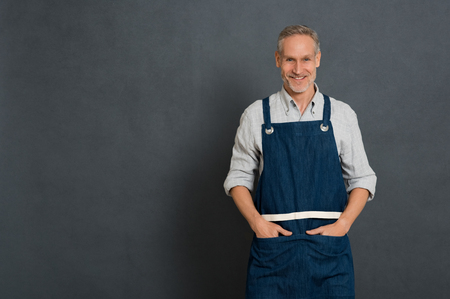 Mature small business owner standing and looking at camera isolated on grey wall. Happy man in work clothes with apron standing. Portrait of successful owner smiling with copy space.の写真素材