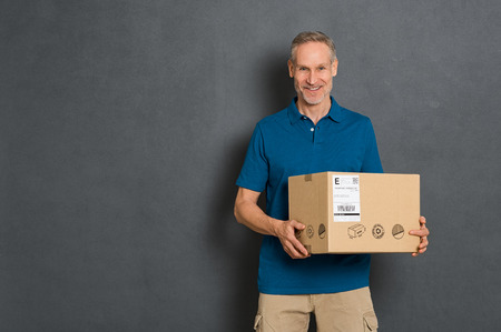 Happy courier holding cardboard box and looking at camera. Smiling delivery man holding and carrying a card box isolated on grey background. Portrait of deliveryman holding parcel with copy space isolated.の写真素材