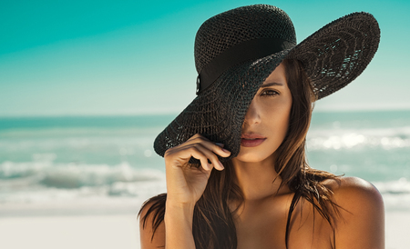 Portrait of fashion young woman posing against sea wearing a black straw hat. Sensual girl covering half face with hat on beach with copy space. Attractive girl looking at camera with sea in background.の写真素材