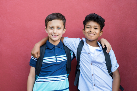 Best children friends standing with hand on shoulder against red background. Happy smiling classmates standing together on red wall after school. Portrait of multiethnic schoolboys enjoying friendship.の写真素材