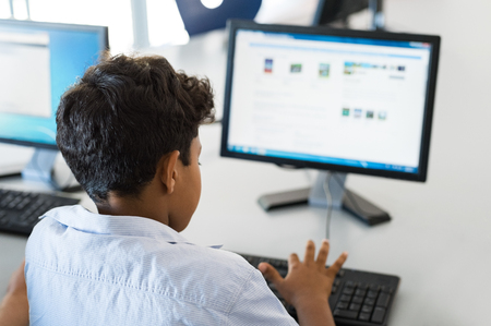 Rear view of multiethnic boy using desktop computer in school library. Back view of mixed race child working in classroom with computer at elementary school. Young arab schoolboy typing on keyboard.の写真素材