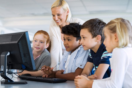 Teacher helping multiethnic children at computer terminal in primary school. Smiling school children looking at computer screen with teacher in library. Happy mature teacher and elementary students using desktop pc in classroom.の写真素材
