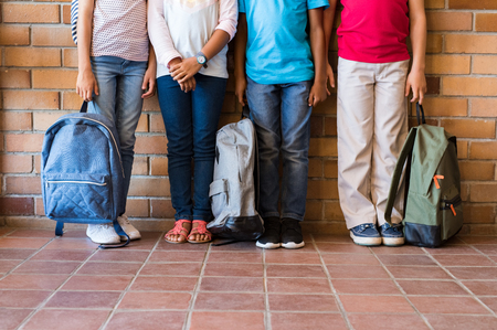 Low section of elementary students standing outside class with backpacks. Legs of four boys and girls leaning in a row. Four multiethnic school children before the start of the lessons.の写真素材