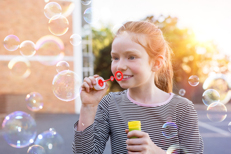 Portrait of lovely little girl with red hair blowing soap bubbles at park. Portrait of cute girl blowing soap bubbles outside the school. Happy female child playing with soap bubble at playground.の写真素材