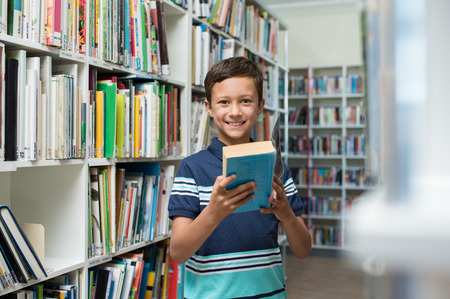 Happy smiling boy reading a book in the library at school. Portrait of cute child in a bookstore looking at camera. Confident pupil borrow a textbook from the library.の写真素材