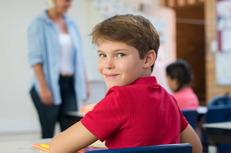 Portrait of smart school boy in classroom with teacher in background. Close up face of schoolchild looking at camera during lesson. Smiling boy looking behind while teacher teaching class.の写真素材