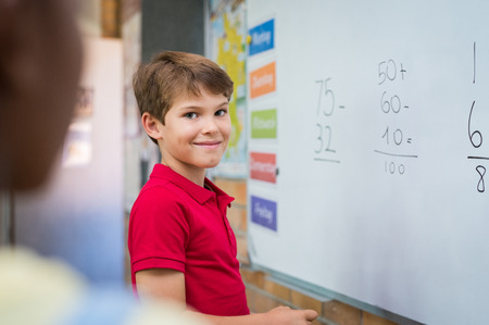 Clever boy doing sums on white board and looking at camera. Smiling schoolboy solving addition and subtraction sum in mathematics lesson. Proud young boy standing with math operation solved. の写真素材