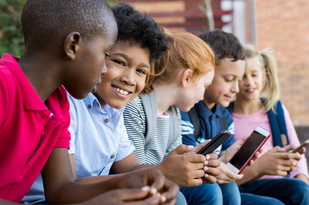 Portrait of smiling male child sitting in a row with other children while using cellphone outdoor. Cute pupils typing on mobile phone at the elementary school during recreation time. Kids holding smartphone.の写真素材