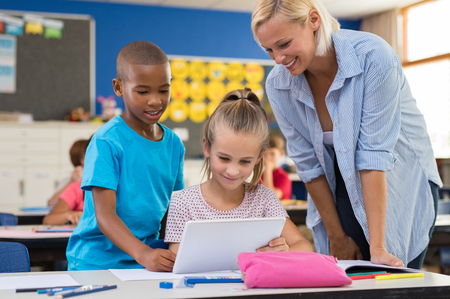 Multiethnic young students using digital tablet in classroom with teacher. Teacher in class with kids using electronic tablet. Teacher teaching how to use electronic device to children at elementary school.の写真素材