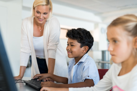 Teacher teaching to elementary class how to use the computer. Happy smiling boy in a computer room learning to use computer. Middle eastern schoolboy in IT class surfing the net at laptop.の写真素材
