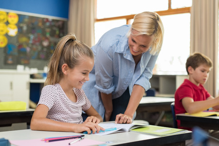 Happy teacher helping young girl with study in classroom. Teacher teaching to smiling schoolgirl at elementary school. Diligent child sitting at school desk with tutor.の写真素材