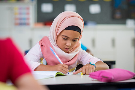 Little muslim girl wearing hijab and doing homework in class. Young arab schoolgirl in chador writing during exam in classroom. Pretty elementary child writing notes during lesson at school.