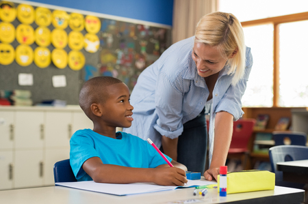 Teacher helping young african boy with homework. Happy teacher helping her student at elementary school. Portrait of woman teaching to schoolchild in classroom. Elementary school teacher helping students.の写真素材