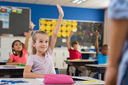 Multiethnic group of young school children raising their hands to answer a question posed by the teacher. Group of elementary kids sitting in classroom and raising hands. Clever girl raising hand knowing the answer.の写真素材