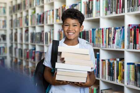 Middle eastern boy holding a stack of books against multi colored bookshelf in library.Â Portrait of happy arab schoolboy with bagpack holding a heap of books at school. Happy young indian child holding books.の写真素材