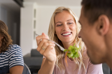 Happy smiling girl feeding salad to her boyfriend. Cheerful young woman giving healthy food salad to man for lunch. Romantic couple and friends enjoying healthy food together at home.の写真素材