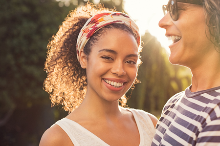Cheerful african woman with hair band standing with boyfriend and looking at camera. Young multiethnic couple enjoying sunset at park. Portrait of beautiful black girl with toothy smile feeling good.の写真素材
