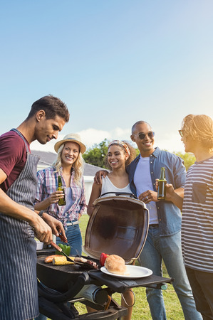 Group of multiethnic young friends enjoying barbecue in the backyard. Happy men and beautiful women talking and grilling meat. Smiling guys and girls having a good time at outdoor party during sunset and drinking beers.の写真素材