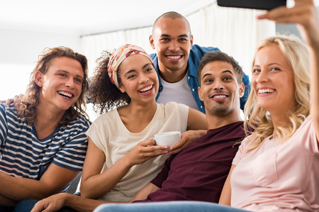 Group of multiethnic friends taking selfie at home. Smiling men and young women with smart phone taking selfie while sitting on couch. Group of funny boys and happy girls taking photo with smartphone. の写真素材