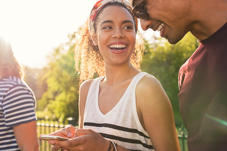Cheerful african woman holding smartphone and looking her boyfriend. Happy young couple reading message on mobile phone and laughing against sunset.の写真素材
