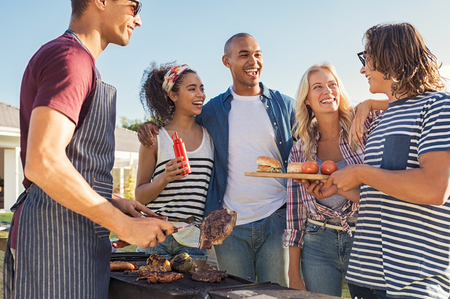 Young multiethnic friends having fun grilling meat enjoying bbq party. Group of happy guys and girls cooking and eating at barbecue dinner outdoor.の写真素材